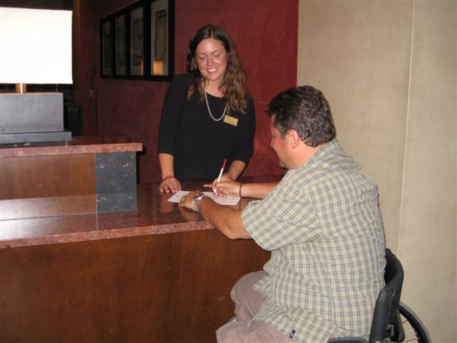 A man in a wheelchair sits at a hotel front desk, signing paperwork while a hotel staff member watches