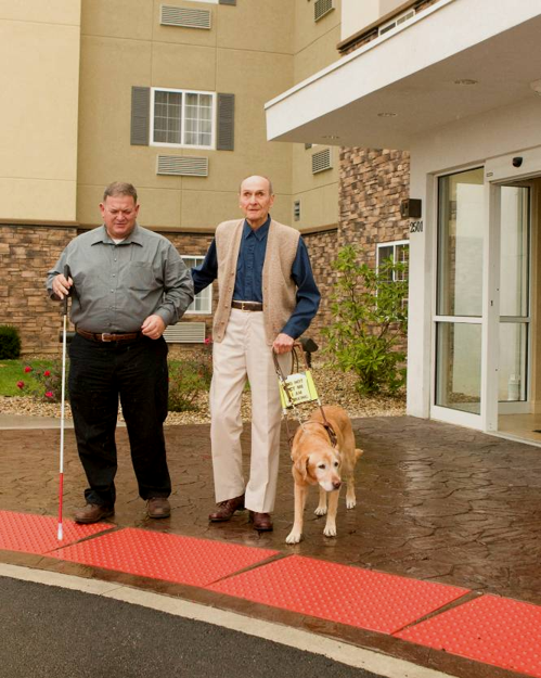 Two men who are blind standing in front of a hotel entrance, left holding a white cane, right with a guide dog